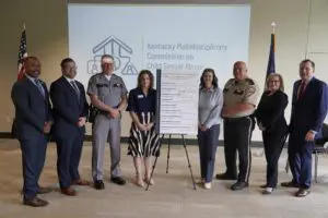 Image of 8 people standing around a sign with a logo in the back for the KY Multidisciplinary Commission on Child Sexual Abuse.