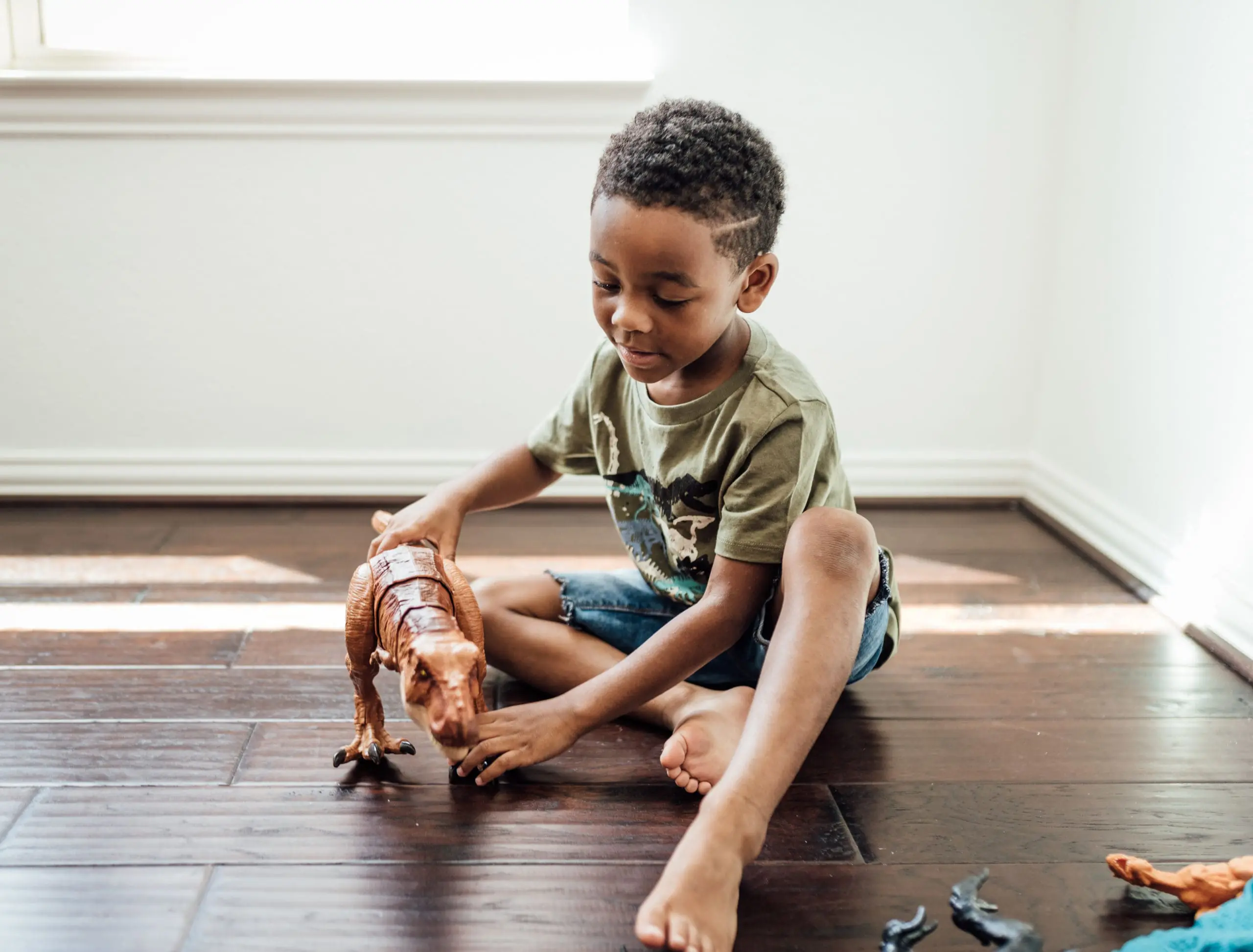 A child sits on a wooden floor, joyfully playing with a large dinosaur toy.