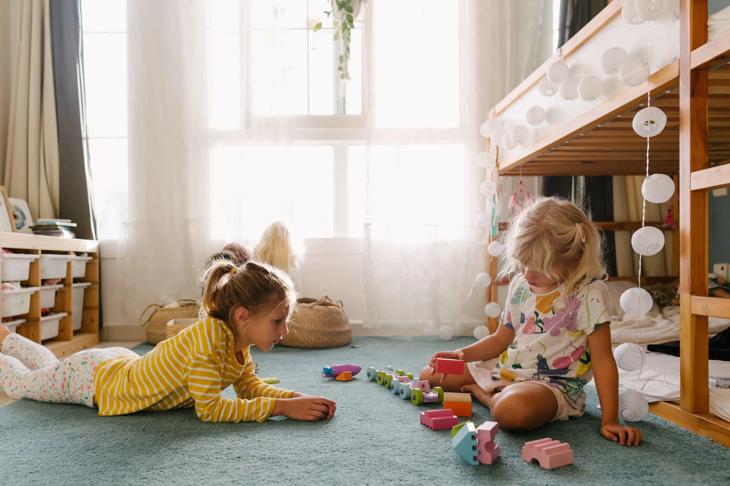 Two children are playing together on the floor in a sunlit room, surrounded by toys.
